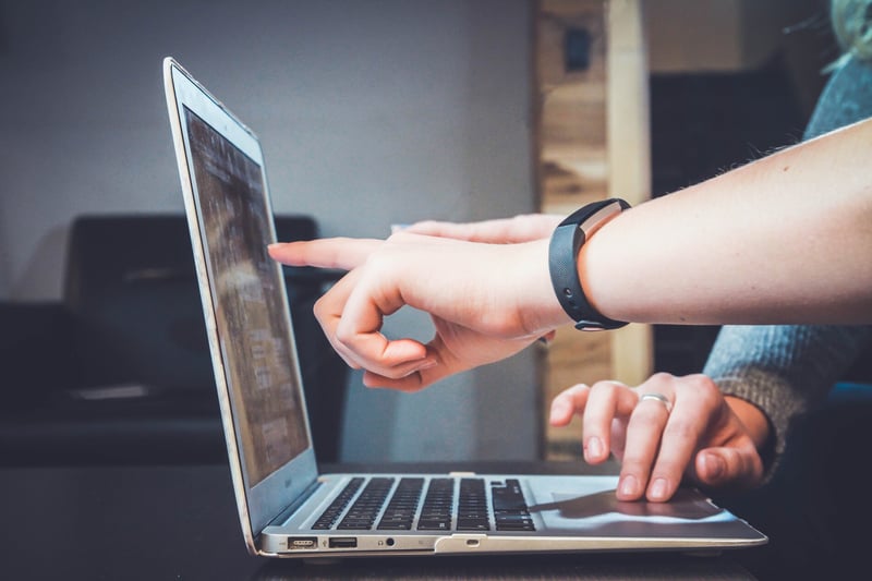 Hände die auf den Bildschirm eines Laptops zeigen, der auf dem Tisch steht, andere Hand ist auf dem Touchpad