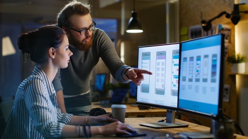 Ein Business Man und eine Business Woman unterhalten sich vor zwei PC-Bildschirmen