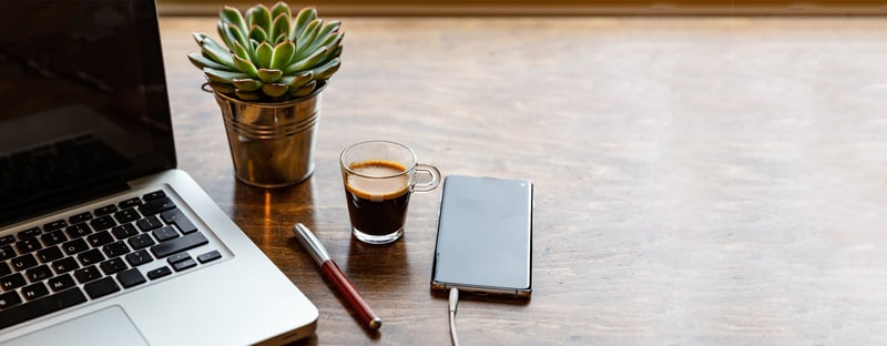 Auf einem Tisch liegen ein Notizblock, ein Kugelschreiber und ein Handy, daneben steht eine Tasse Kaffee
